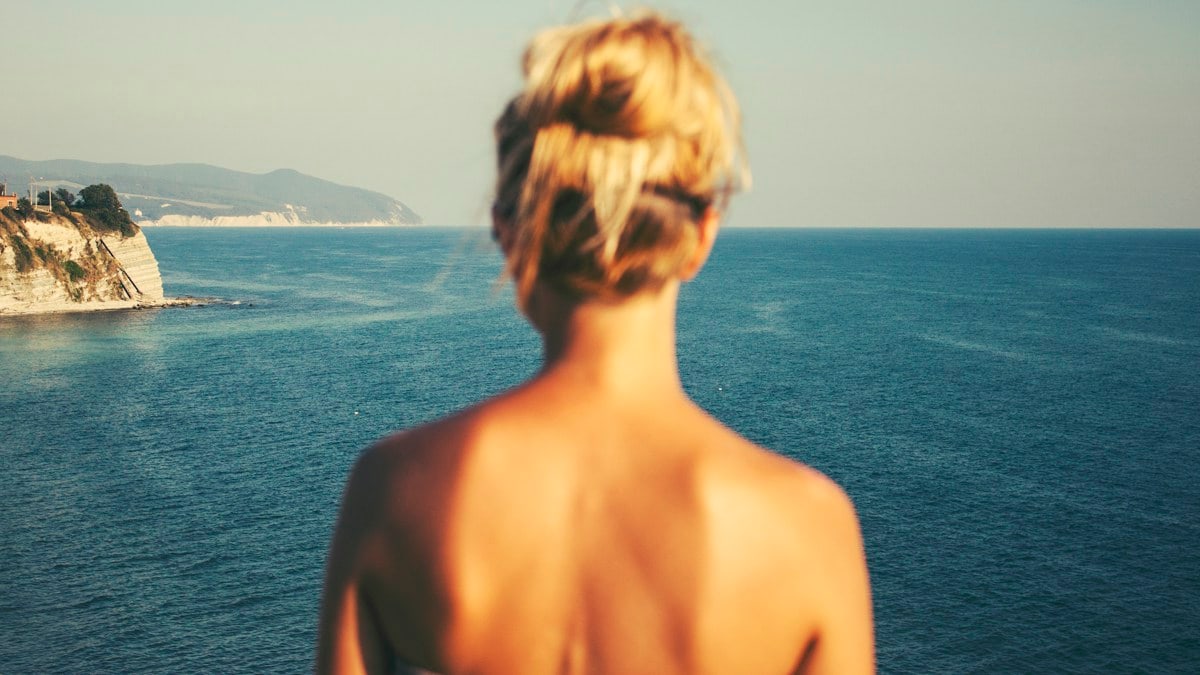 topless woman standing facing blue sea under white sky