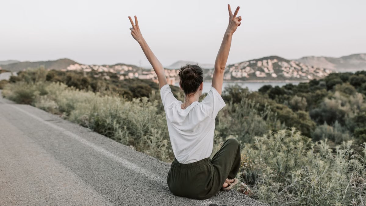 Woman on a roadside in Antalya raises peace sign, embracing summer with mountain views and tranquility.